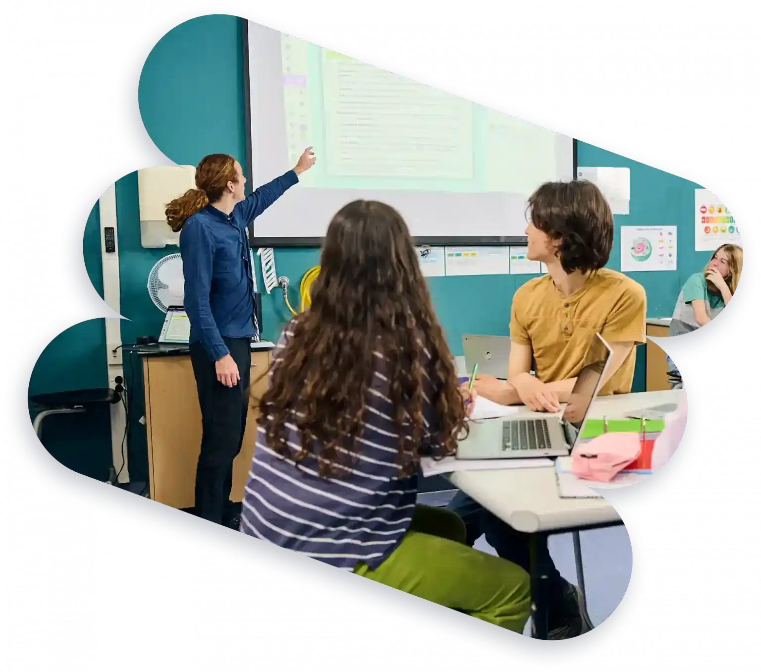 A teacher stands at the front of a classroom pointing at a projected screen, while three students sit at desks facing her, listening and looking at laptops and notebooks.