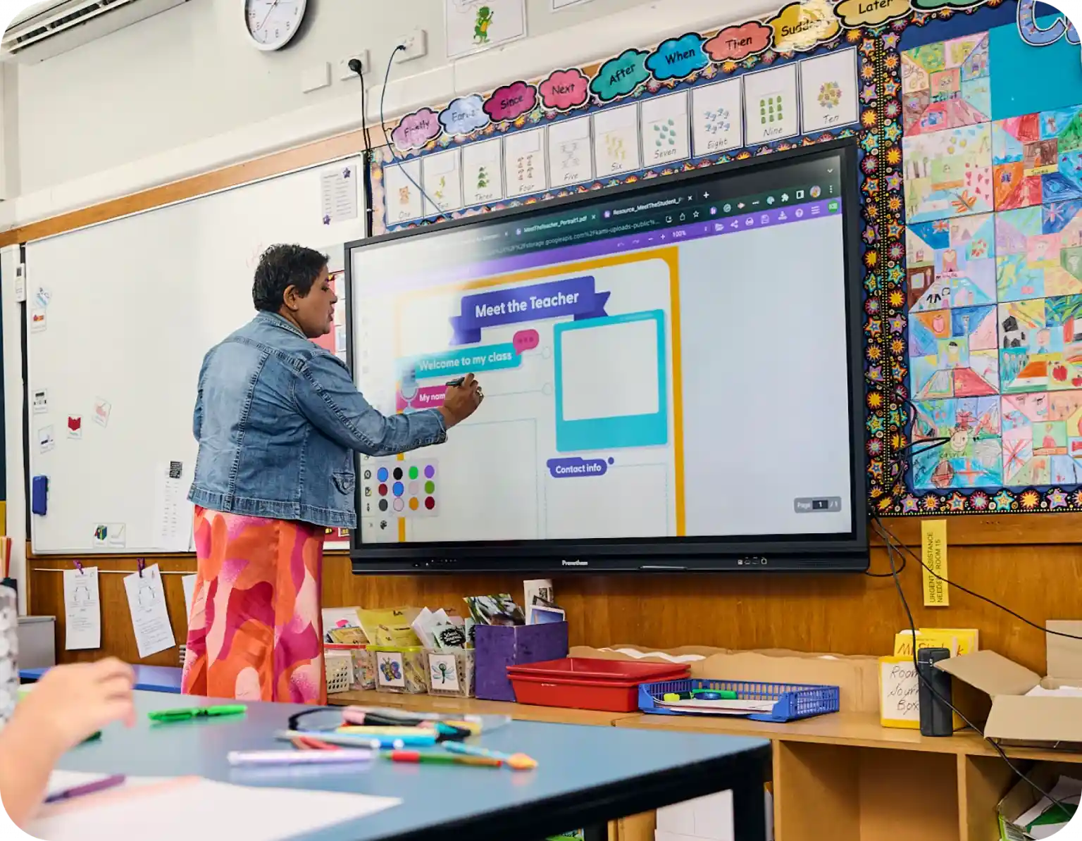 A teacher in a colorful skirt and denim jacket uses a large touchscreen display in a classroom decorated with student artwork and educational posters. The screen shows a Meet the Teacher slide.