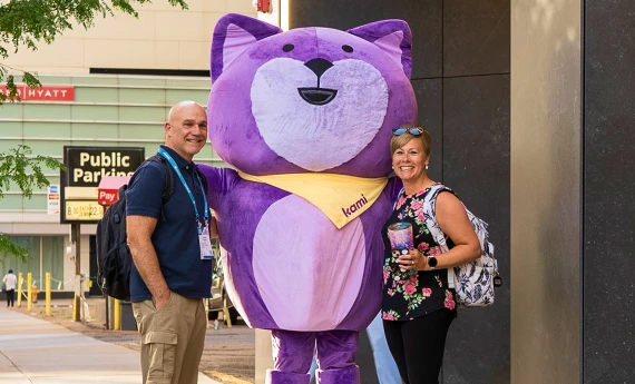 A man and woman smiling while standing with the Kami mascot at an educational conference.
