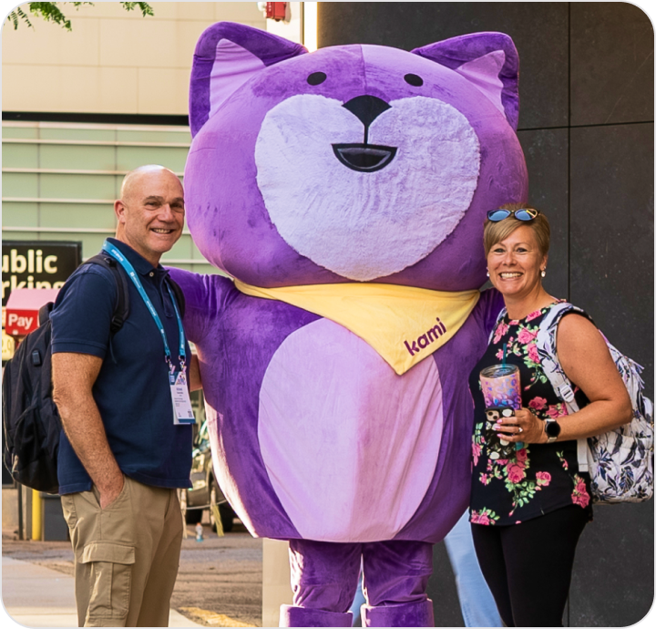 Two people posing beside a large purple dog mascot wearing a ‘Kami’ sash, in a public venue; a pink heart graphic appears on the right.