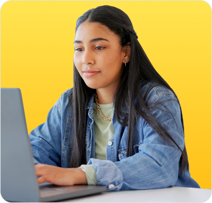 Student with long dark hair wearing a denim jacket, sitting at a laptop against a bright yellow background.