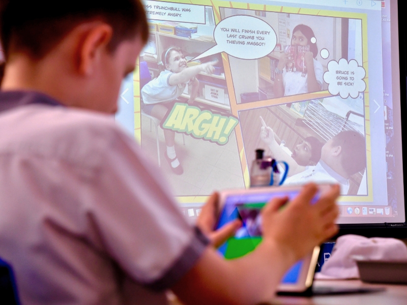 A child works on a tablet in front of a projector screen displaying a Book Creator comic strip with speech bubbles.