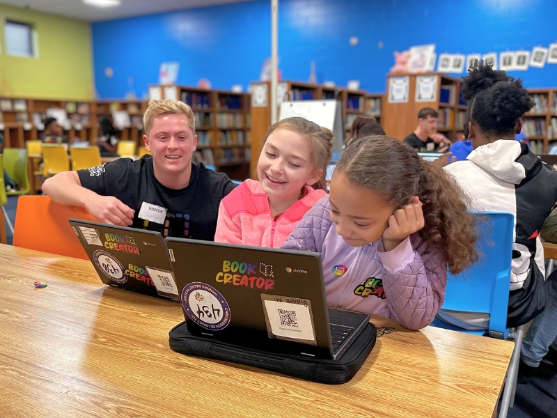 A man smiles at two girls as they work on laptops in a library. The laptops have colorful stickers, including one that says Book Creator.