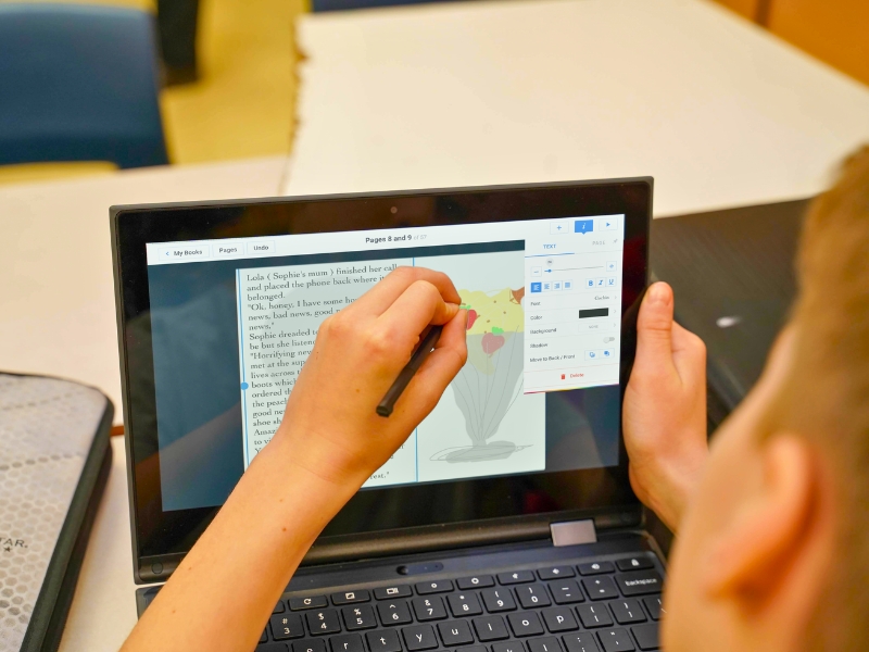 A student uses a stylus to draw an ice cream sundae in Book Creator on a laptop screen displaying a document with handwritten notes.