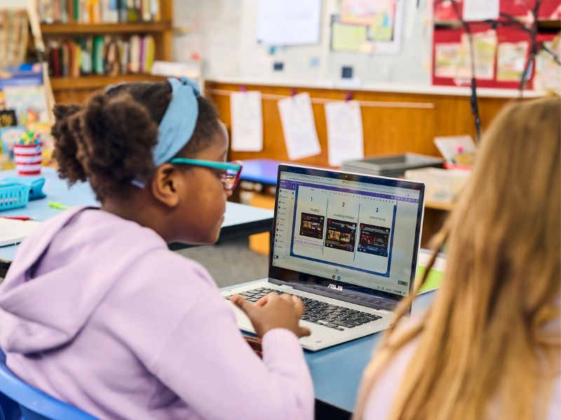 A student wearing glasses and a purple hoodie looks at a laptop screen displaying three images on Kami App. Bookshelves are visible in the background of the classroom