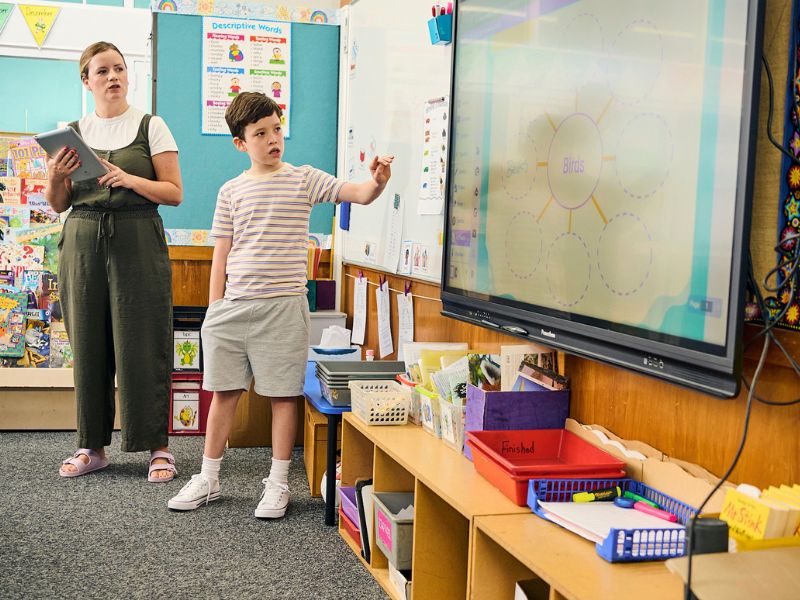 A teacher holding a tablet stands next to a student pointing at a digital whiteboard displaying Kami App in a classroom