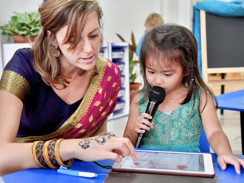 A teacher in a sari with henna on her hand and bangles points at a tablet screen while a child holds a microphone to their mouth to record her voice in Book Creator.