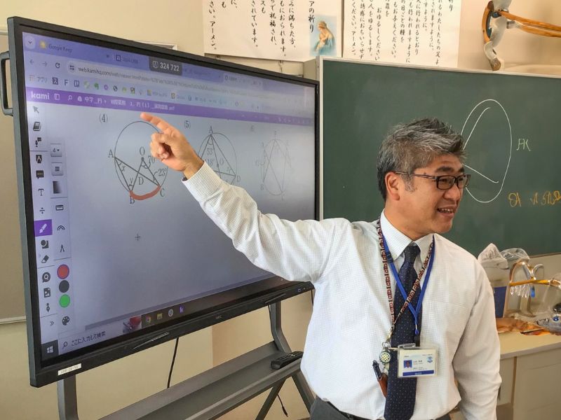 A teacher points to a diagram of a circle in the Kami App on a smartboard while standing in a classroom