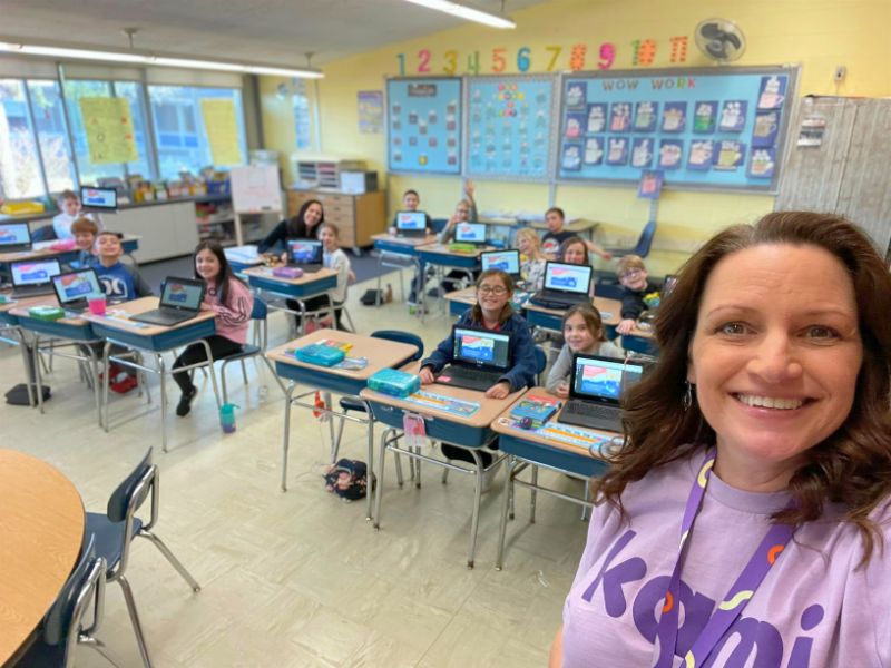 A teacher smiles at the camera in the foreground, with a classroom of students at desks with laptops behind her