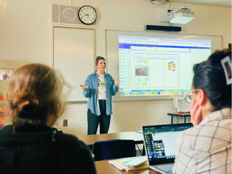 A teacher stands in front of a projector screen showing Kami App in a classroom, gesturing with her hands as she speaks to students seated at desks