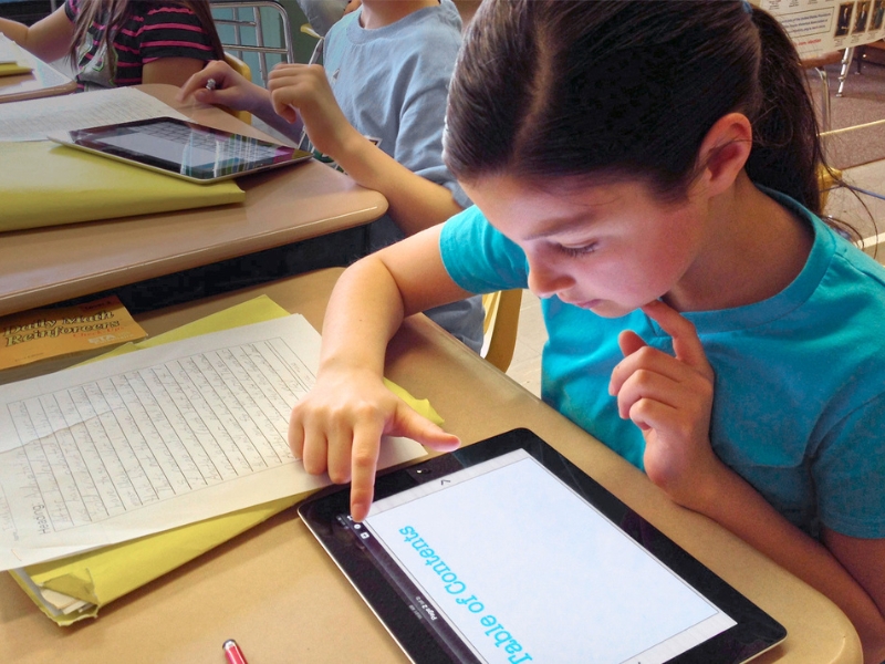 A young person is focused on a tablet screen whilst using Book Creator, displaying the words Table of Contents. She is sitting at a classroom desk with books and papers.