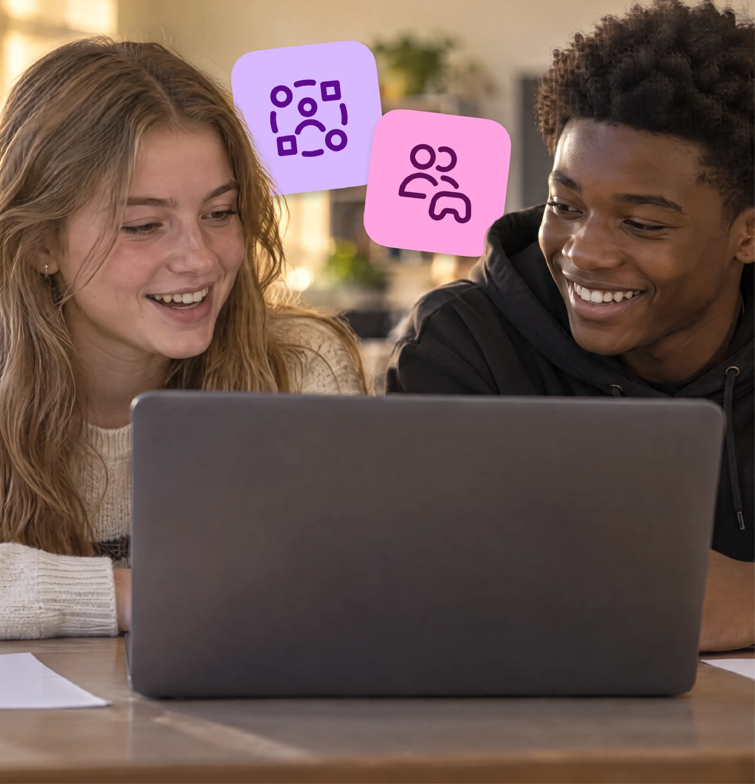 Two high school students smile and talk as they work together on a shared laptop.